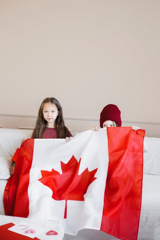 2 little girls holding the flag cloth of Canada. One of them is hiding from camera and the other is smiling at it.