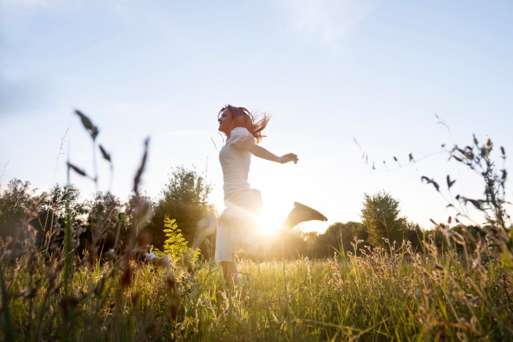 A smiling girl , dressed in a white dress with puffy half sleeves, runs with her arms open in a field with lush green grass as sun shines. 